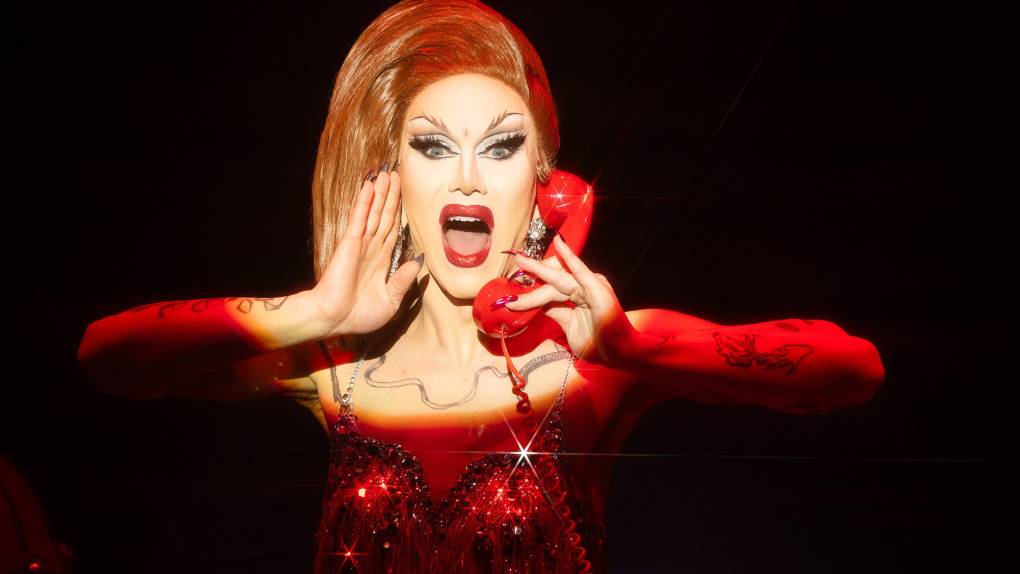 A drag queen with large red hair and a red dress holds a red telephone with a red cord wearing red lipstick