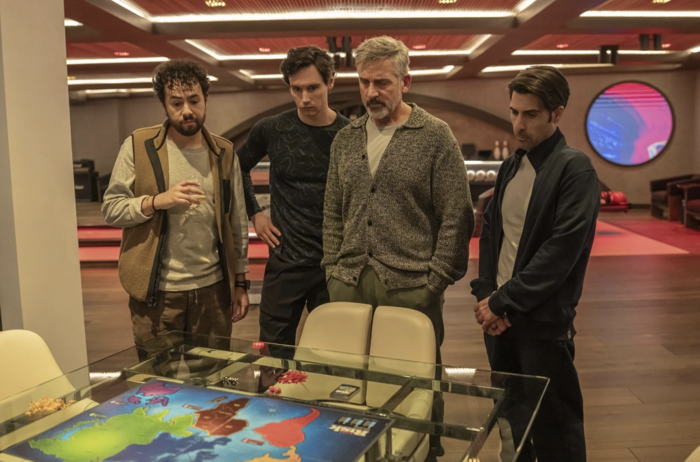 Four men in casual clothes stand in a large windowless room, staring at a board game that features a map of the world.