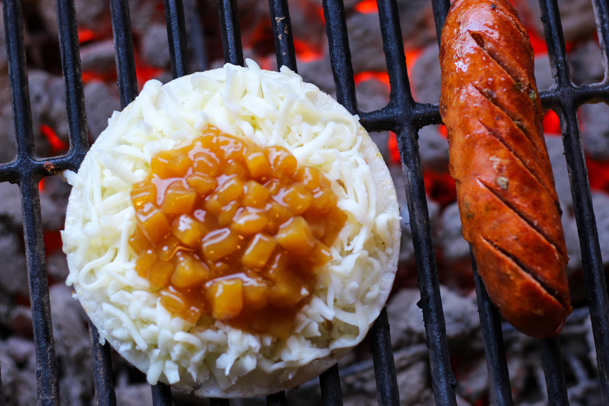 An arepa topped with shredded cheese and pinapples cooking over a charcoal grill, alongside a sausage link.
