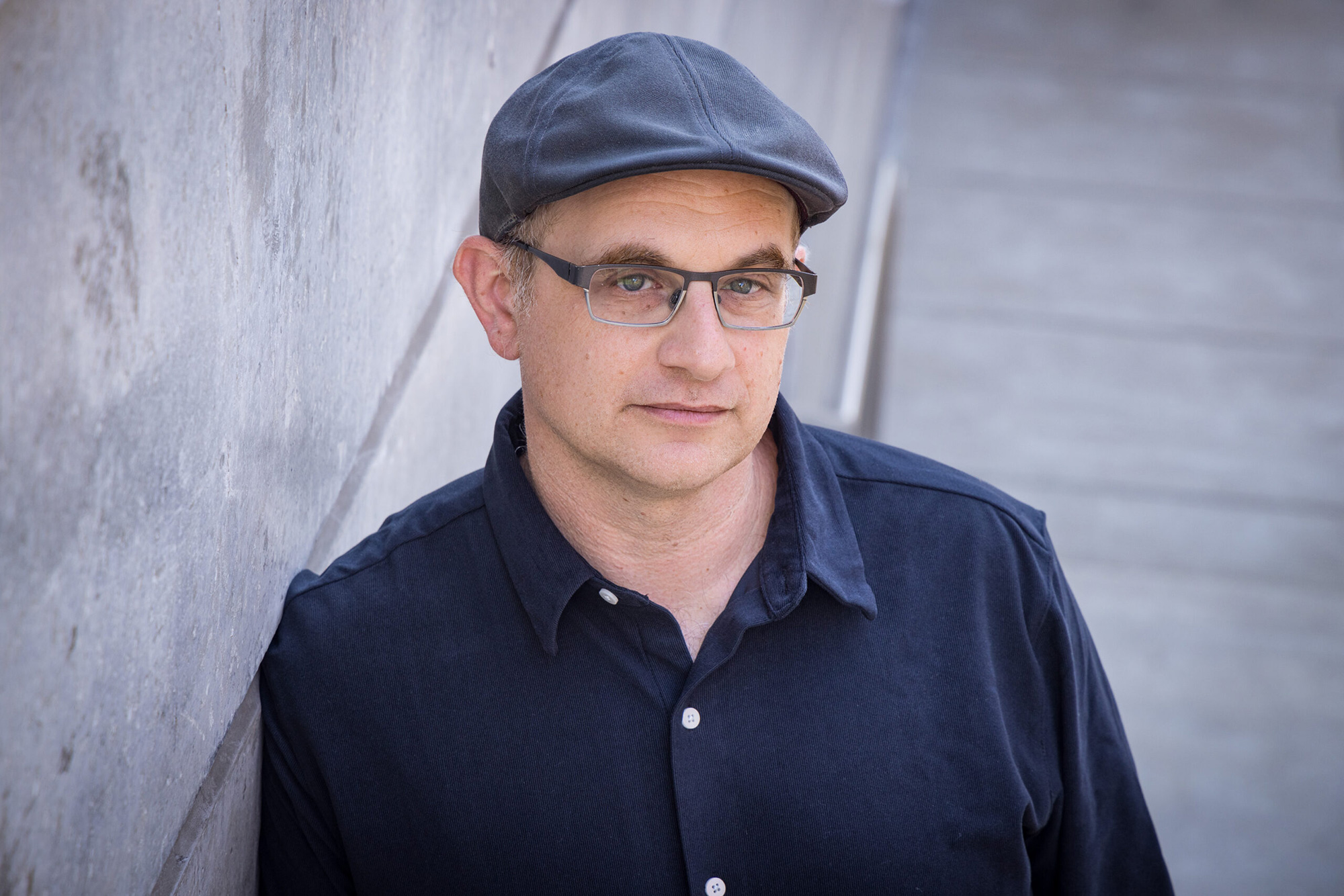 A man in glasses and a newsboy cap looks upward at the camera